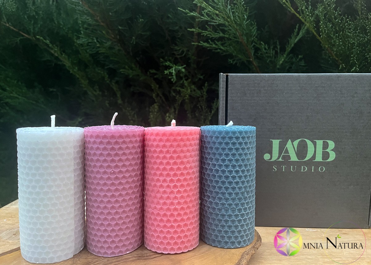 JaobStudio-Candele di Cera d’Api Colorate Presentazione delle candele naturali Jaobstudio. Sono allineate e disposte su un supporto di legno grezzo naturale in un'atmosfera bucolica in esterno. Dietro le candele si vede la confezione di cartone grigia con il marchio dell'azienda al centro della stessa. Le candele sono di colore bianco, rosa lilla, rosa, blu.