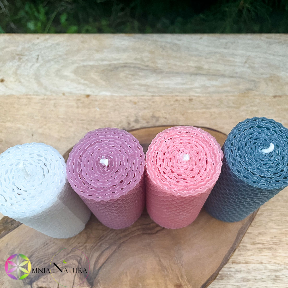 JaobStudio-Candele di Cera d’Api Colorate Candele di cera d'api disposte una accanto all'altra per mostrare la loro caratteristica manifattura arrotolata. Sono fotografate in un esterno e come base sono su posizionate su due basi di legno naturali. bianca, rosalilla, rosa confetto e azzurro polvere. Si vede in sfondo del verde del prato.