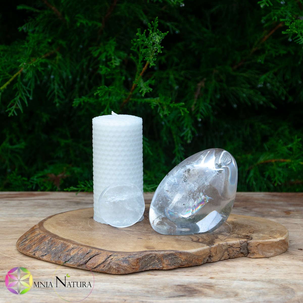 Candela di cera d’api Snow Ball – JaobStudio In una magica giornata di pioggia la candela bianca è presentata al centro con due pietre lavorate di meraviglioso quarzo ialino trasparente. Una pietra piatta burattata appoggiata in verticale davanti e una grande forma libera con arcobaleno interno visibile a destra. Evocativo il ginepro verde sullo sfondo e molto bello anche la parte di legno grezzo su cui si poggia il tutto. bianco, verde, legno, arcobaleno, di grande impatto.