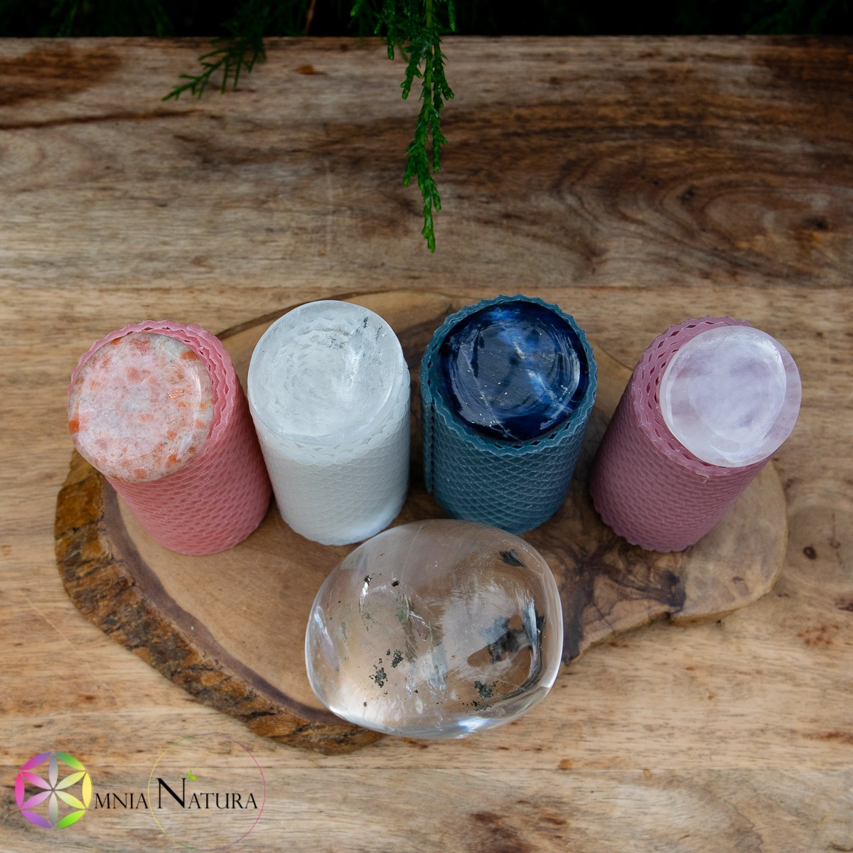JaobStudio-Candele di Cera d’Api Colorate Le candele naturali JaobStudio sono fotografate dall'alto in un contesto naturale esterno. In un'evocativa giornata di pioggia sono disposte in fila su un supporto di legno grezzo. In questo caso sono 4, rosa, bianca, blu polvere, rosa lilla, Sopra ognuna è posizionata una pietra piatta burattata differente: Pietra di Sole Naturale, Quarzo Ialino trasparente, Sodalite e quarzo rosa. Di fronte alle candele c'è una splendida forma libera di quarzo ialino con inclusioni.