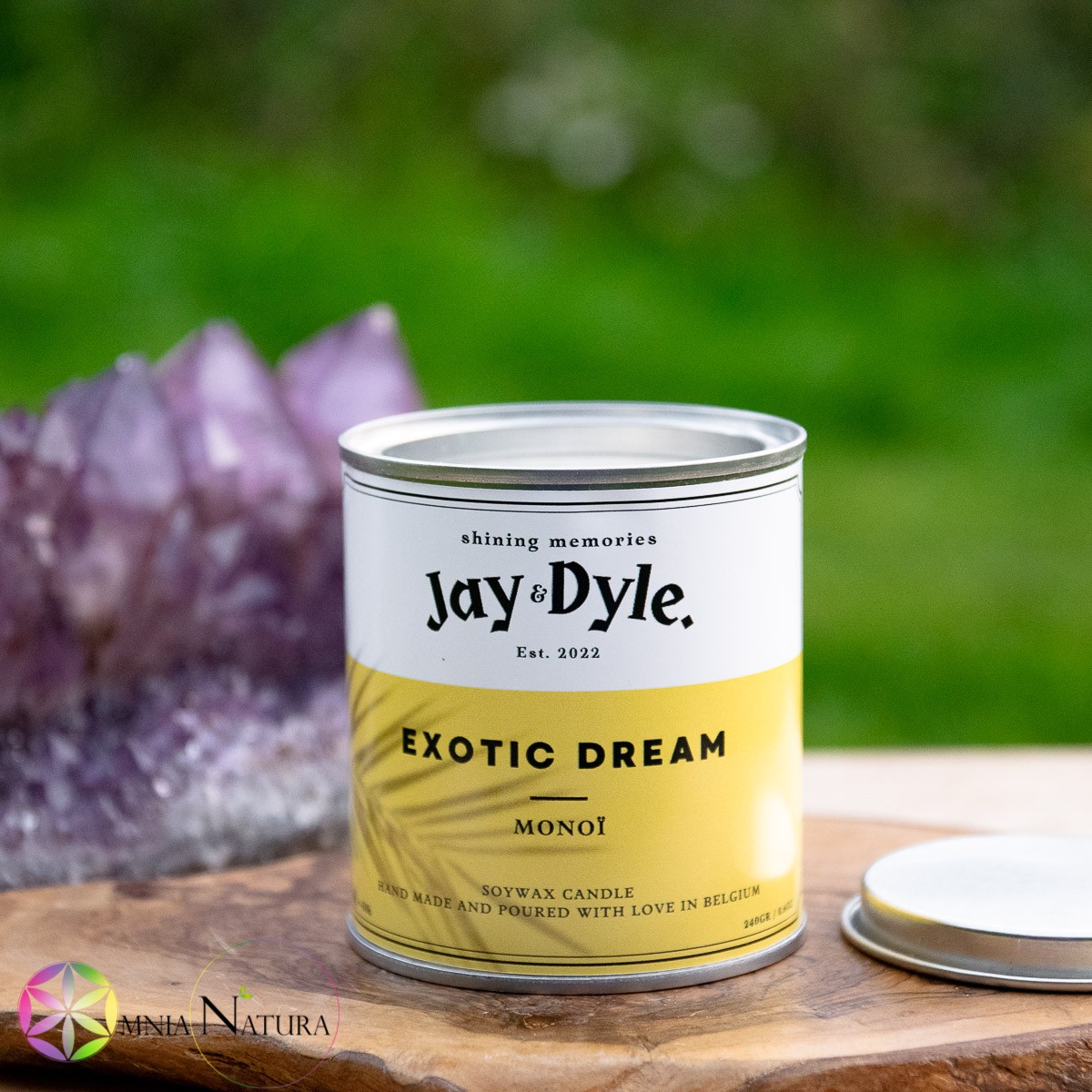 Jay And Dyle-Exotic Dream-Monoi Candela in Soia 100% naturale in contenitore di metallo colorato e originale, in stile simile ad un barattolino di vernice, con coperchio aperto. La candela è appoggiata su un tagliere di legno e ha come sfondo il verde di un giardino e un'ametista dai grandi cristalli.