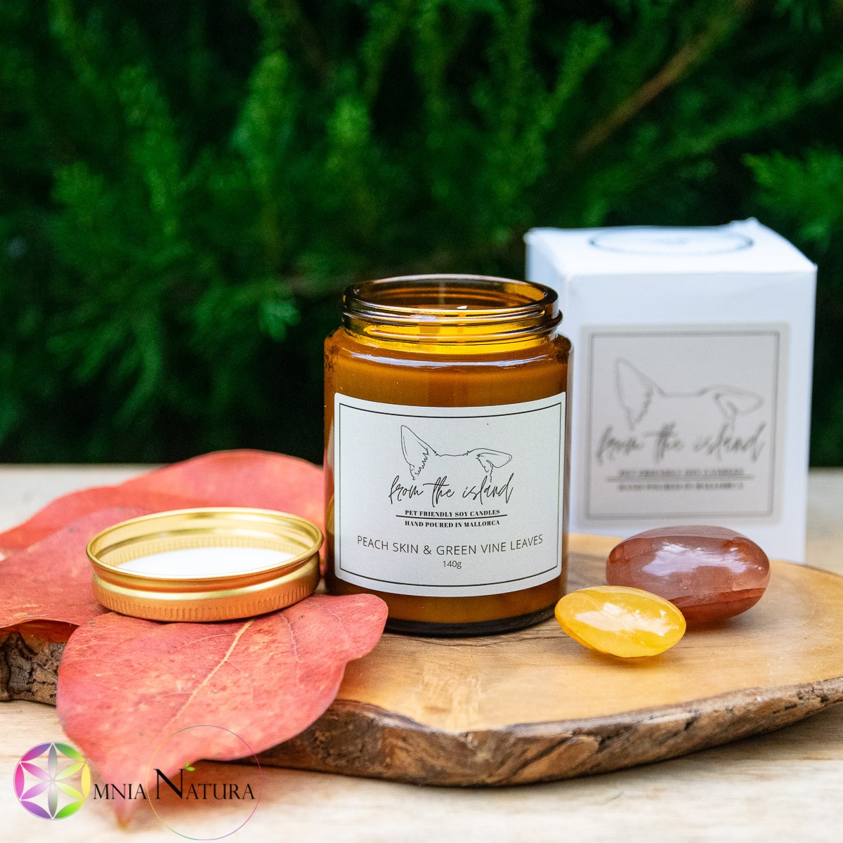 Peach Skin and Green Vine Leaves “From the island” in questa foto viene presentato il vasetto di vetro ambrato in cui è versata a mano la candela naturale pet friendly di FROM THE ISLAND. Il vasetto è aperto e il tappo dorato di metallo è appoggiato vicino, adagiato su una foglia autunnale di colori caldi. il tutto è disposto su un tagliere di legno grezzo e accanto alla candela sono disposte due pietre burattate per esaltare la bellezza del tutto. Una pietra è un pebble di quarzo ematoide rosso e l'altra una lens di quarzo ematoide giallo. Sullo sfondo la confezione in cartone di FROM THE ISLAND e la natura vera e viva con i suo verde ad icorniciare il tutto.